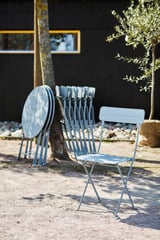 Ett set med blå SUNDSÖ klappstolar på en uteplats, perfekt för små utrymmen. En är öppen, andra hopvikta, lutade mot ett träd.