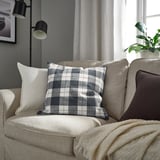 Beige soffa med SVAMPFLY rutig kudde, kuddar, växt och lampa.