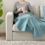 Person som sitter på en beige soffa, håller en vit mugg, täckt med en ljusblå fleecefilt.