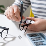 En person använder en precisionsskruvmejsel från TRIXIG-setet för att fixa glasögon. Den magnetiska bitslådan och handtaget är synliga på bordet.