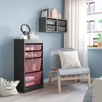 TROFAST svart enhet med rosa korgar, hörlurar, böcker och vit väska. Hög, nära stol, blå vägg.