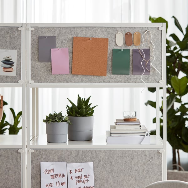 En modern förvaring med hyllor som visar växter, böcker och anteckningar. Ett dubbelsidigt skåp, en whiteboard och en anslagstavla i kork för organisation.