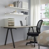 Beige TROTTEN skrivbord med svarta ben, under vita hyllor, svart stol, fönster, växt.