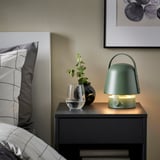 Grön VAPPEBY-lampa på nattduksbordet, med växt och kudde. Bärbar, mjuk glöd.