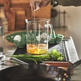 Person som blandar vätska i en genomskinlig måttkanna med ingredienser och örter på en köksbänk.
