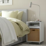 Modern sovrumsscen med ljusgrå stoppad säng, beige sängkläder och ett grått och rotting sidobord med lampa och vaser.