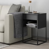 Svart VIKHAMMER sidobord med lådor bredvid beige soffa, som rymmer böcker.