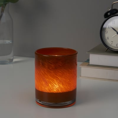 Cylindrisk ljusstake i glas med orange virvlar, som avger varmt ljus, bredvid böcker och en klocka.