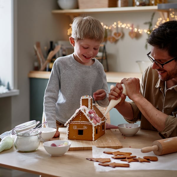 Man och barn dekorerar pepparkakshus med glasyr, omgivna av godis och skålar på ett träbord.