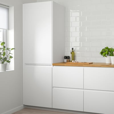 Modernt vitt VOXTORP-kök med högt kylskåp, eleganta vita skåp, bänkskiva i trä och krukväxter.