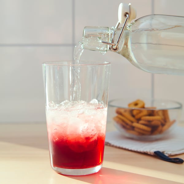 Clear jar pours liquid into glass with red drink, beside pretzels.