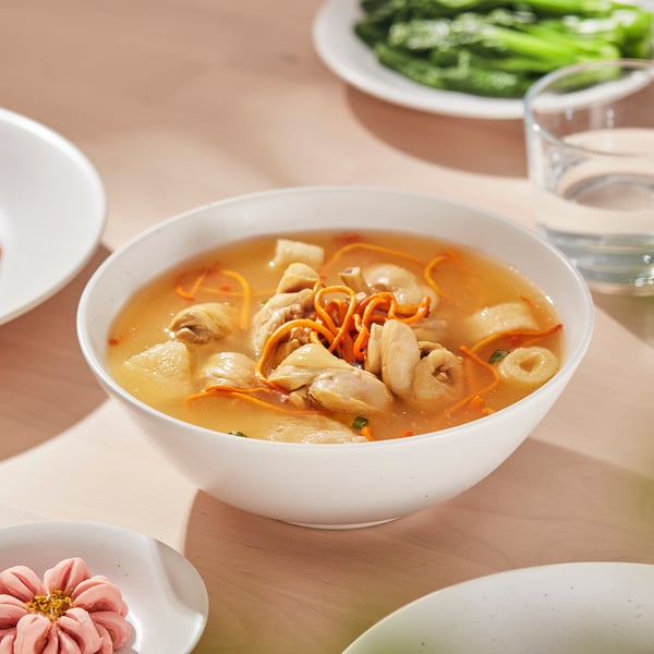 A white porcelain bowl filled with chicken and noodle soup sits on a wooden table surrounded by other dishes and a glass of water.