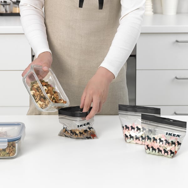 Person holding clear container pouring mixed nuts into re-sealable ikea ISTAD bag on counter top, showing organization and food storage.