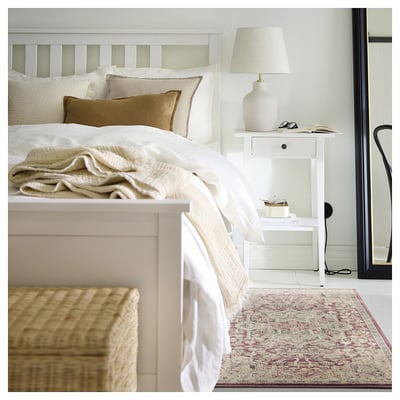 Bedroom with white bed, cream pillows, beige blankets, white bedside table, lamp, and colourful rug.