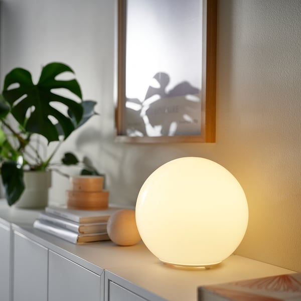Small round KAJPLATS lamp on table, beside books & plant, lit in warm light.