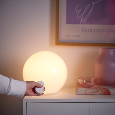 Hand holding remote control for smart lamp on white side table.