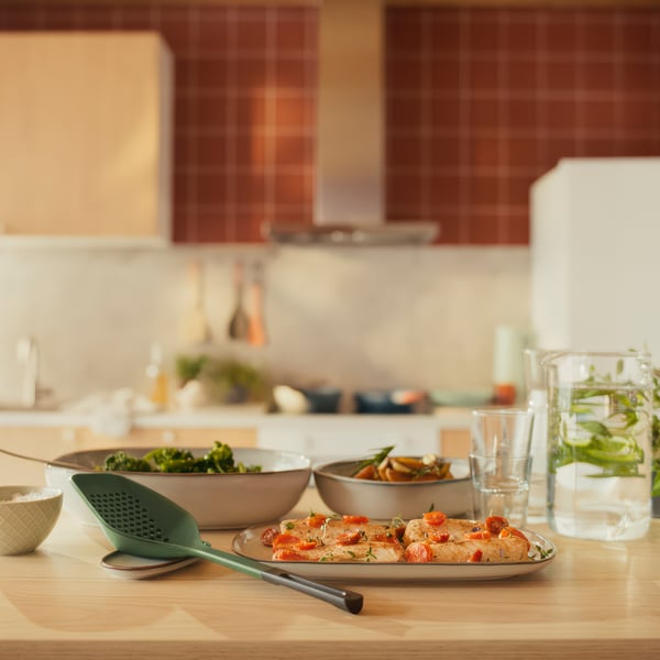A kitchen scene with a green KNORRHANE utensil on a wooden table, holding roasted vegetables, next to bowls and water glasses.