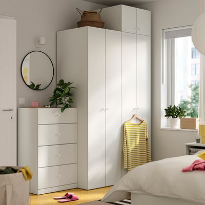 Bedroom with white LASTARE wardrobe and chest, yellow and pink accents, greenery, and a round mirror.