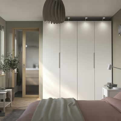 Modern bedroom with white PAX wardrobe, pink bed linens, black pendant lamp, olive tree, and wooden mirror.