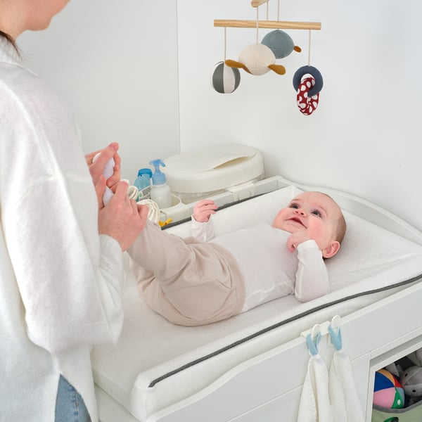 A person tends to a baby on a changing table, with diapers and creams nearby. The table rear shelves for storage and is designed for baby care.
