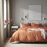 Sodobna glavna spalnica z veliko posteljo, oranžno-rjavo odejo, resnično vidno umetnostjo, elegantnim dekorjem, naravno svetlobo, elegantno nočno omarico in visokimi cvetovi.
