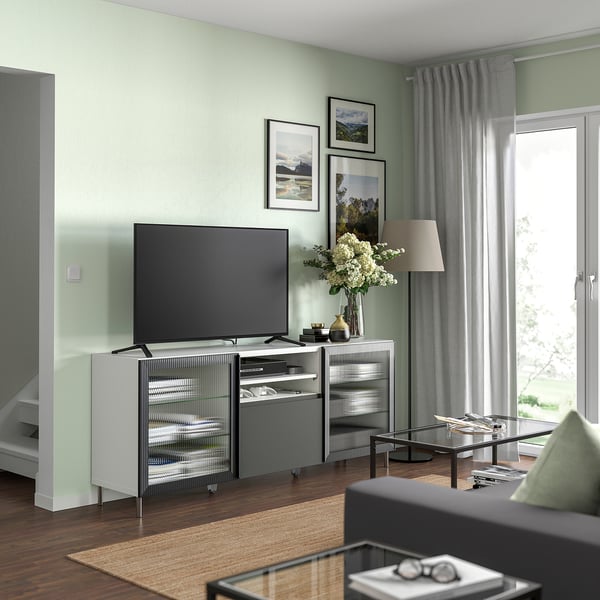 Sodobna dnevna soba z belo TV klopjo BESTÅ, TV z ravnim zaslonom, sivimi zavesami, lesenimi tlemi, preprogo, svetilko, vazo in uokvirjenimi fotografijami.