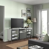 Sodobna dnevna soba z belo TV klopjo BESTÅ, TV z ravnim zaslonom, sivimi zavesami, lesenimi tlemi, preprogo, svetilko, vazo in uokvirjenimi fotografijami.