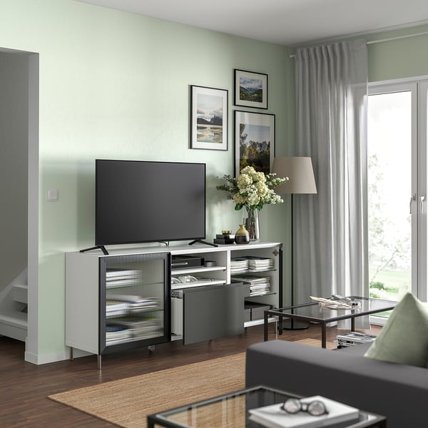 Sodobna dnevna soba s TV klopjo BESTÅ v beli in črni barvi. Velika televizija z ravnim zaslonom na vrhu, steklena omara spodaj, sive zavese in lesena tla.