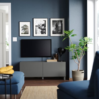 Sodobna dnevna soba: temne stene, TV klop, ploski zaslon, svetilka, rastlina, modri kavč, rumena blazina, fotografije, lesena tla, preproga.