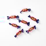 Osem rdeče zavitih bonbonov DUMLE z modrimi konci je raztresenih po beli površini. DUMLE gladka čokolada in mehko žvečilno karamelno nadev.