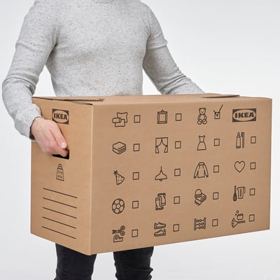Oseba, ki drži veliko kartonsko premikajočo se škatlo z oznako ikea z izrezanimi ročaji in različnimi simboli, ki označujejo vsebino.