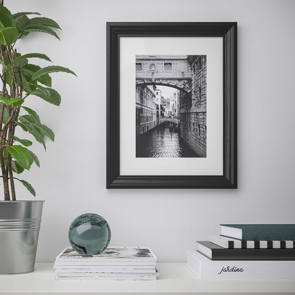 Črno uokvirjena fotografija na beli steni, kanal z mostom in dekorativni predmeti spodaj.