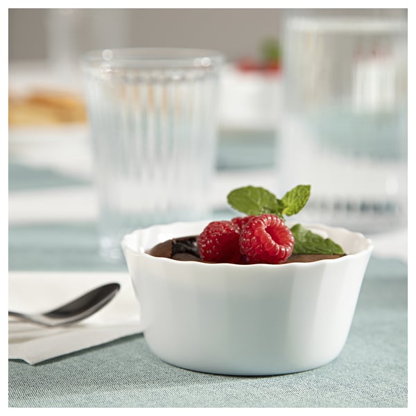 Bela keramična posoda, napolnjena s čokoladnim moussejem, posuta z malinami in listi mete, postavljena na moder prt poleg kozarca.