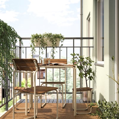 Balkon z akacijevim lesom FURUÖN in belo kovinsko jedilno garnituro; Vključuje mizo, stole in podnožje. Obkrožen z lončnicami, ki ustvarjajo prijeten prostor.