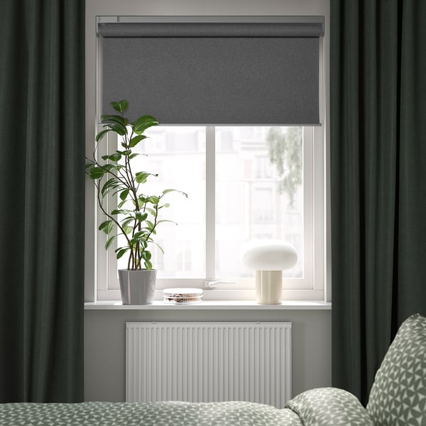 Sodobna spalnica s sivo roletno, temne zavese, beli radiator, blazine, svetilka, visoka rastlina.