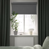 Sodobna spalnica s sivo roletno, temne zavese, beli radiator, blazine, svetilka, visoka rastlina.