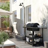 Dvorišče s črnim žarom na oglje GRILLSKÄR, nerjaveče jeklo, pravokotno, na keramični terasi ob sivi hišni steni.