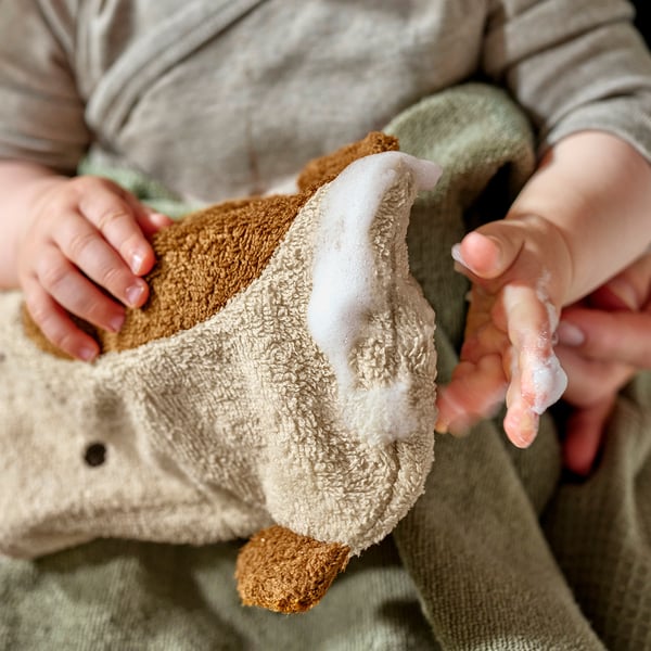 Otrok, ki drži bež rokavico v obliki žirafe GULDVÄVARE. Mehak, organski bombaž z vezenim obrazom, kot nalašč za nežno čiščenje.