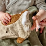 Otrok, ki drži bež rokavico v obliki žirafe GULDVÄVARE. Mehak, organski bombaž z vezenim obrazom, kot nalašč za nežno čiščenje.