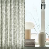 Sodobna soba z zeleno-belimi geometrijskimi zavesami HÅRKÖRVEL, elegantno okensko polico, svetilko, knjigami in minimalističnim dekorjem.