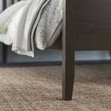 Temno rjavi okvir postelje IDANÄS z lepo zloženo belo odejo s cvetličnim vzorcem. Okvir je eleganten, koničaste noge in trden lesen zaključek.
