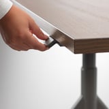 Oseba, ki prilagaja višino mize IDÅSEN z ročajem. Miza ima nastavljivo višino in upravljanje kablov.