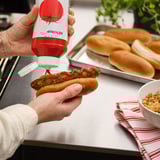 Oseba stisne rdeči KEČAP na hotdog, ki ga drži v roki.