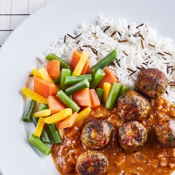 Krožnik rastlinskih mesnih kroglic v curry omaki s pisano parjeno zelenjavo in divjim rižem.