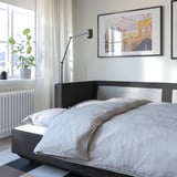 Sodobna spalnica s sivim raztegljivim kavčem LANDSKRONA, uokvirjenim mostom, radiatorjem in rastlino ob oknu.