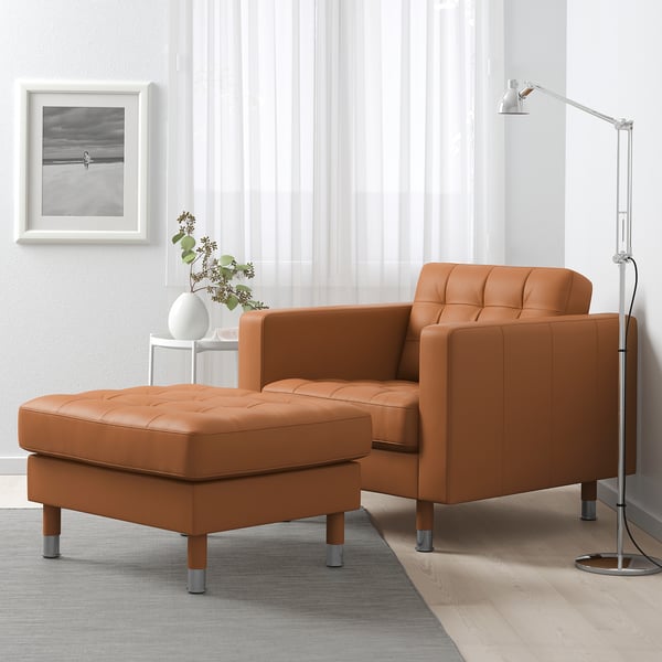 Sodobna dnevna soba z rjavim usnjenim foteljem IKEA in otomanom, sivo preprogo, talno svetilko in stensko umetnostjo.