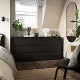 Sodobna spalnica s črno-rjavo komodo MALM v bližini okna, eleganten dizajn, ogledalo, svetilka in rastlina.