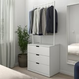 Minimalistična spalnica z belo NORDLI 3-predalno skrinjo pod ograjo za oblačila, zelena rastlina zraven med posteljo in oknom.