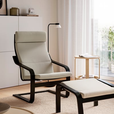 Minimalistični bivalni prostor s POÄNG naslanjačem in podnožnikom iz bež korduroja, črnega lesenega okvirja, poleg stranske omarice z lučjo ob oknu.