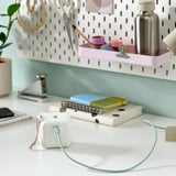 Bela miza z zelenim kablom USB-c, priključenim na majhno napravo, zvezke in rastlino. Pegboard s pisarniškimi potrebščinami v ozadju.